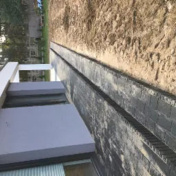 Dwa pasy ułożonej, ciemnej kostki brukowej oddzielone rzędem wystających kostek obrzeżowych, biegnące wzdłuż ściany budynku z szarą elewacją i oknem, na tle piaszczystego podłoża.