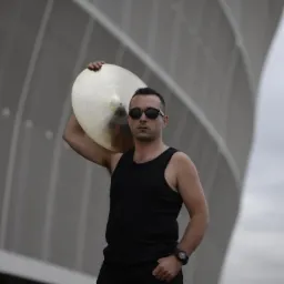 Mężczyzna w czarnej koszulce na ramiączkach i okularach przeciwsłonecznych trzymający na ramieniu talerz perkusyjny na tle nowoczesnej architektury.