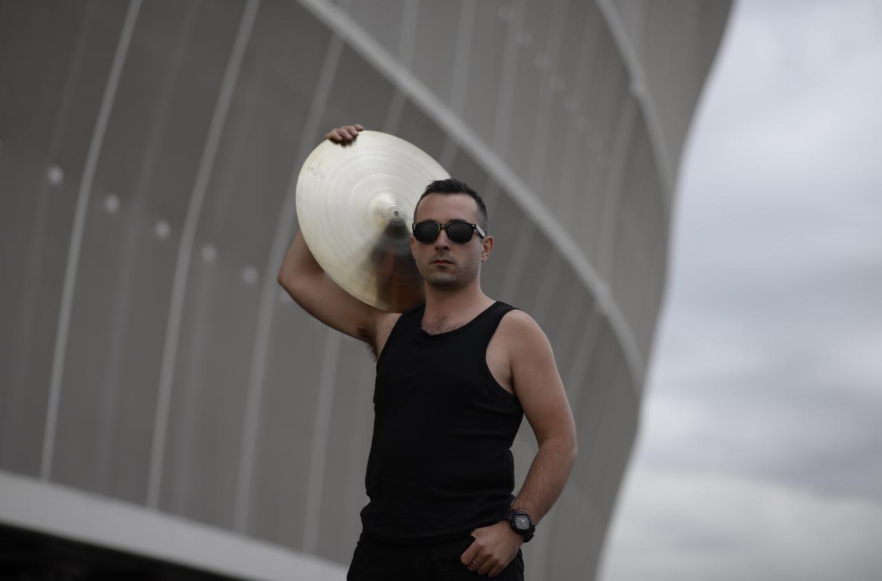 Mężczyzna w czarnej koszulce na ramiączkach i okularach przeciwsłonecznych trzymający na ramieniu talerz perkusyjny na tle nowoczesnej architektury.