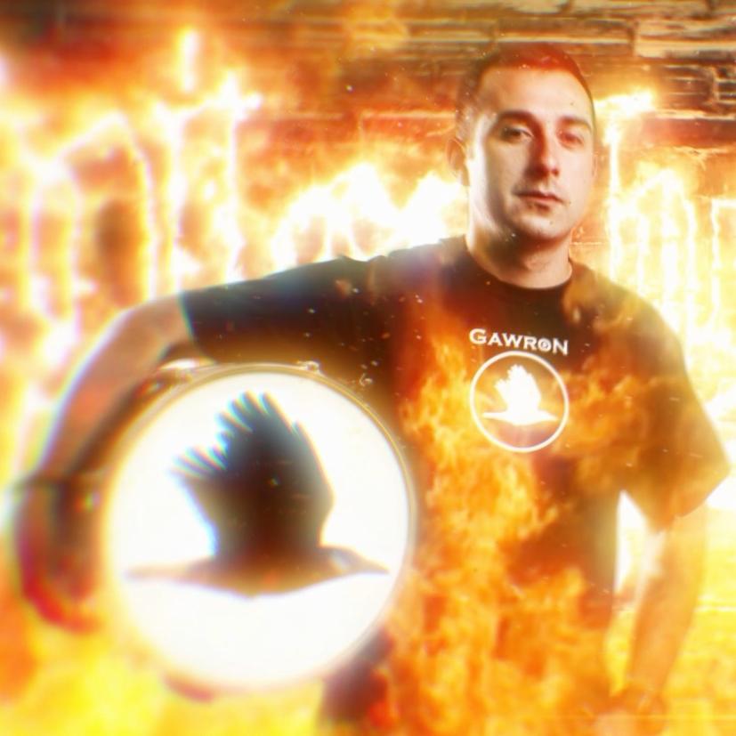 Mężczyzna w czarnej koszulce z logo trzymający tamburyn z motywem ptaka na tle efektu ognia.