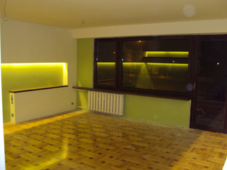 Wnętrze pokoju po remoncie z parkietem ułożonym w jodełkę, świeżo pomalowanymi ścianami w odcieniach zieleni i beżu, z podświetlaną wnęką i grzejnikiem pod oknem.