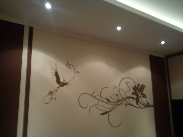 Beżowa ściana z dekoracyjnym, brązowym malunkiem przedstawiającym lecącego żurawia i stylizowaną kompozycję kwiatową, oświetlona punktowymi lampami sufitowymi.