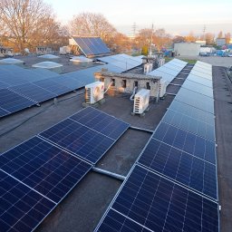 Marek Matras Safe System - Rozległa instalacja paneli fotowoltaicznych na płaskim dachu budynku komercyjnego, z rzędami ciemnych modułów PV, kilkoma klimatyzatorami LG i kolektorem słonecznym w tle, otoczona drzewami bez liści.