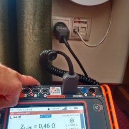 Marek Matras Safe System - Ręka technika obsługuje miernik MPI-540-PV podłączony do gniazdka elektrycznego, wyświetlający wynik pomiaru impedancji pętli zwarcia Z_L-PE = 0,46 Ω. W tle biały router i ciemna zasłona.