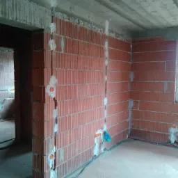 Surowy mur z czerwonej cegły z wyżłobionymi bruzdami wypełnionymi białym gipsem, w kt&oacute;rych poprowadzone są szare kable elektryczne, widoczne pomarańczowe puszki instalacyjne, surowy betonowy sufit.