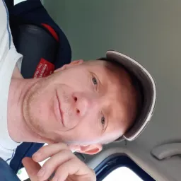 Portret mężczyzny w czapce z daszkiem, wykonany z perspektywy żabiej, w samochodzie, z delikatnym uśmiechem i jasnymi oczami, skierowanymi w stronę obiektywu.