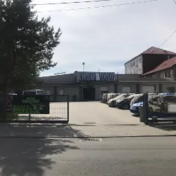 Widok na budynek warsztatu z logo i szyldem firmy oferującej instalacje LPG, z zaparkowanymi samochodami, niektóre przykryte pokrowcami, w tle budynki mieszkalne.