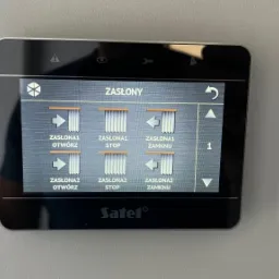 Panel sterowania inteligentnego domu marki Satel, wyświetlający opcje sterowania zasłonami: otwieranie, zamykanie i zatrzymywanie dla zasłony 1 i 2.