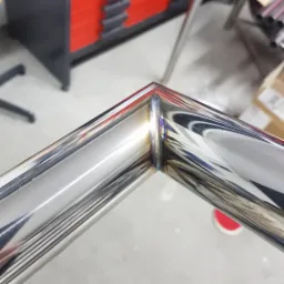 Szczegółowe ujęcie spawu łączącego dwa polerowane, metalowe profile pod kątem prostym, widoczne odbicia otoczenia na powierzchni.