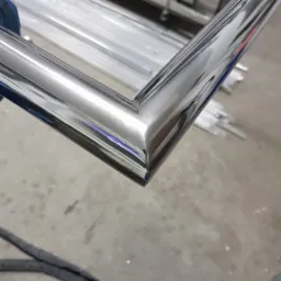 Metalowy profil o lustrzanym połysku, z widocznymi odbiciami otoczenia, leżący na szarym podłożu. W tle widoczne inne elementy metalowe.