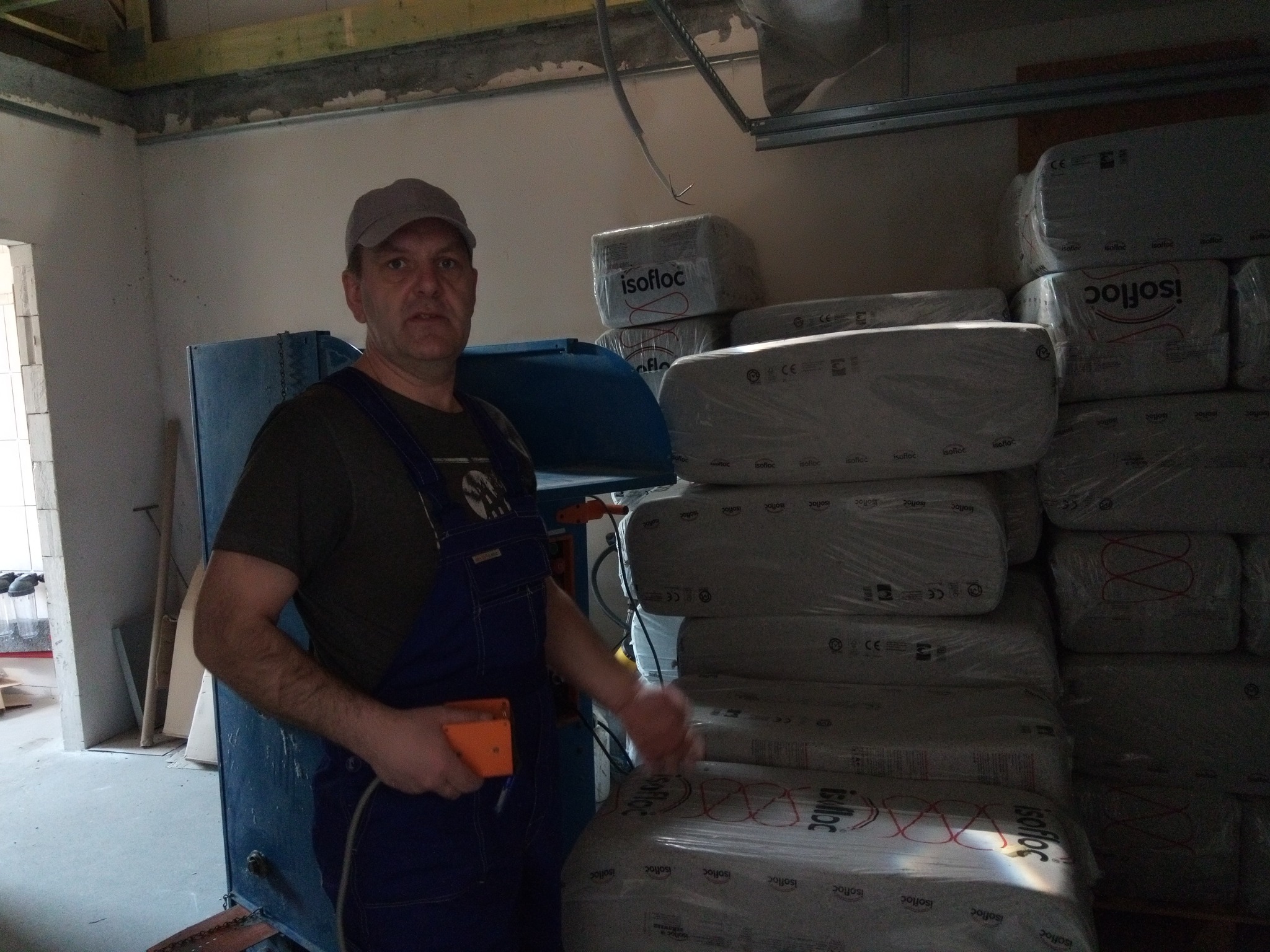Pracownik w kombinezonie roboczym stoi obok maszyny do wdmuchiwania izolacji i sterty zapakowanych worków z materiałem izolacyjnym Isofloc w surowym wnętrzu poddasza.