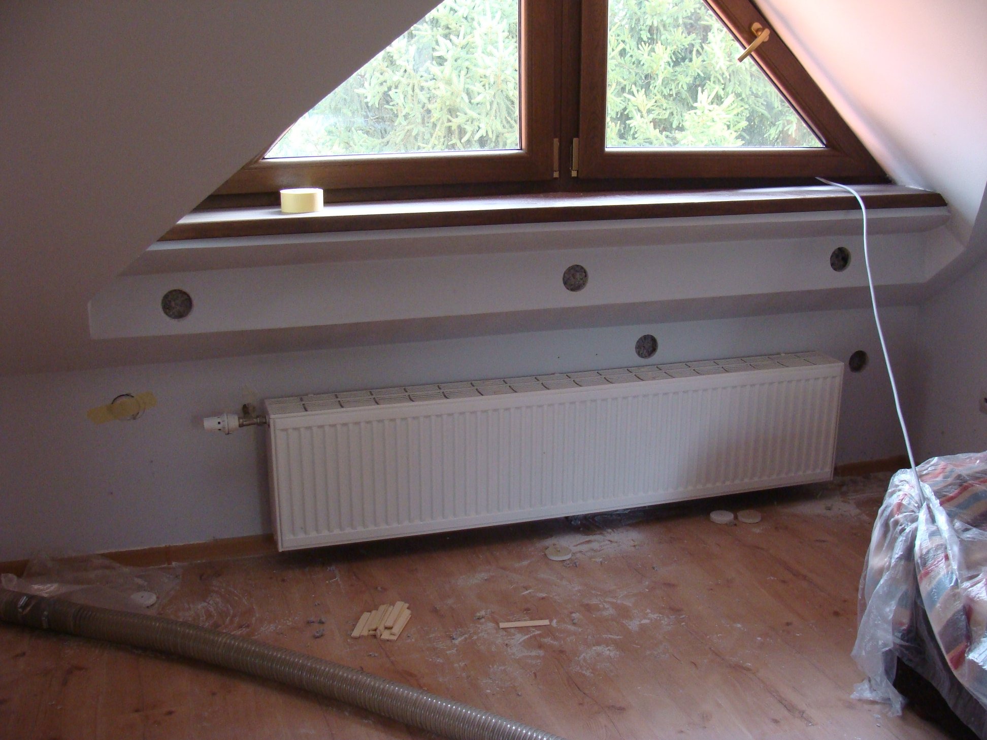 Poddasze z oknem dachowym i zamontowanym grzejnikiem; widoczne otwory w ścianie przygotowane do ocieplenia, rura wentylacyjna i fragment zabezpieczonej folią podłogi.