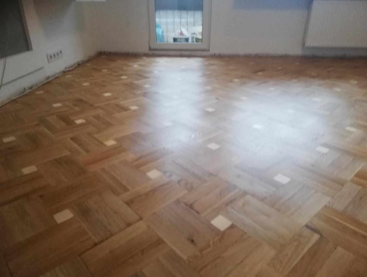 Ułożony parkiet w jodełkę z wstawkami w jasnym kolorze, widok z góry na gotową podłogę w pustym pomieszczeniu z oknem i grzejnikiem.