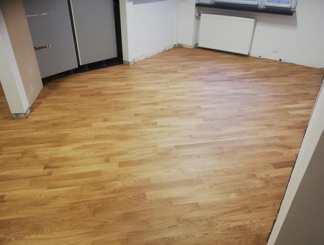 Jasnobrązowe panele podłogowe w pokoju z białymi ścianami i grzejnikiem, widoczne fragmenty drzwi i kolumny, listwy przypodłogowe jeszcze nie zamontowane.