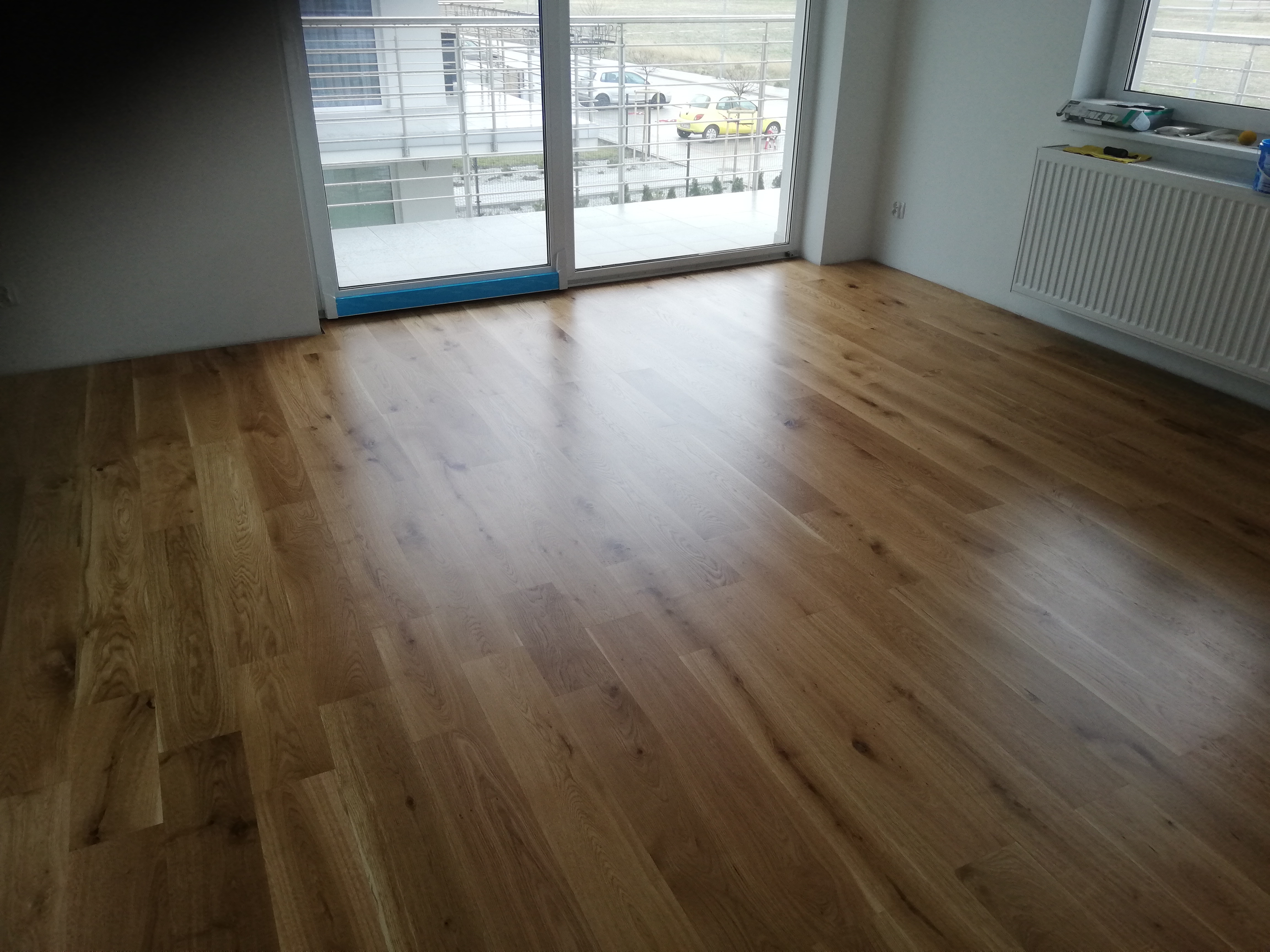Świeżo ułożona, lśniąca podłoga z drewnianych paneli w jasnym odcieniu, widok z perspektywy pokoju na balkon i ulicę przez duże okno balkonowe zabezpieczone niebieską folią ochronną.