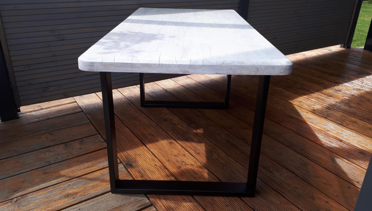 Stół z blatem z bielonego drewna i czarnymi, metalowymi nogami ustawiony na drewnianym tarasie w słoneczny dzień.