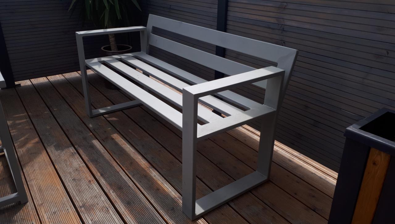 Nowoczesna, metalowa ławka ogrodowa z jasnoszarego profilu kwadratowego, ustawiona na drewnianym tarasie z desek, z widoczną rośliną w donicy w tle.