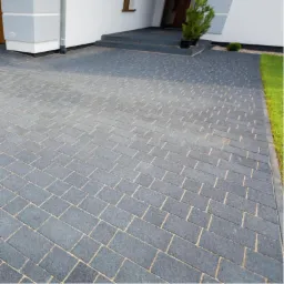 Szara kostka brukowa ułożona na podjeździe domu, z delikatną fugą w kolorze piasku. Widoczne schody wejściowe i fragment trawnika. Ujęcie z niskiej perspektywy.