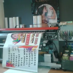 Wielkoformatowa drukarka atramentowa w trakcie drukowania baneru reklamowego z kolorową grafiką, widoczne pojemniki z tuszem i rolki papieru w tle.