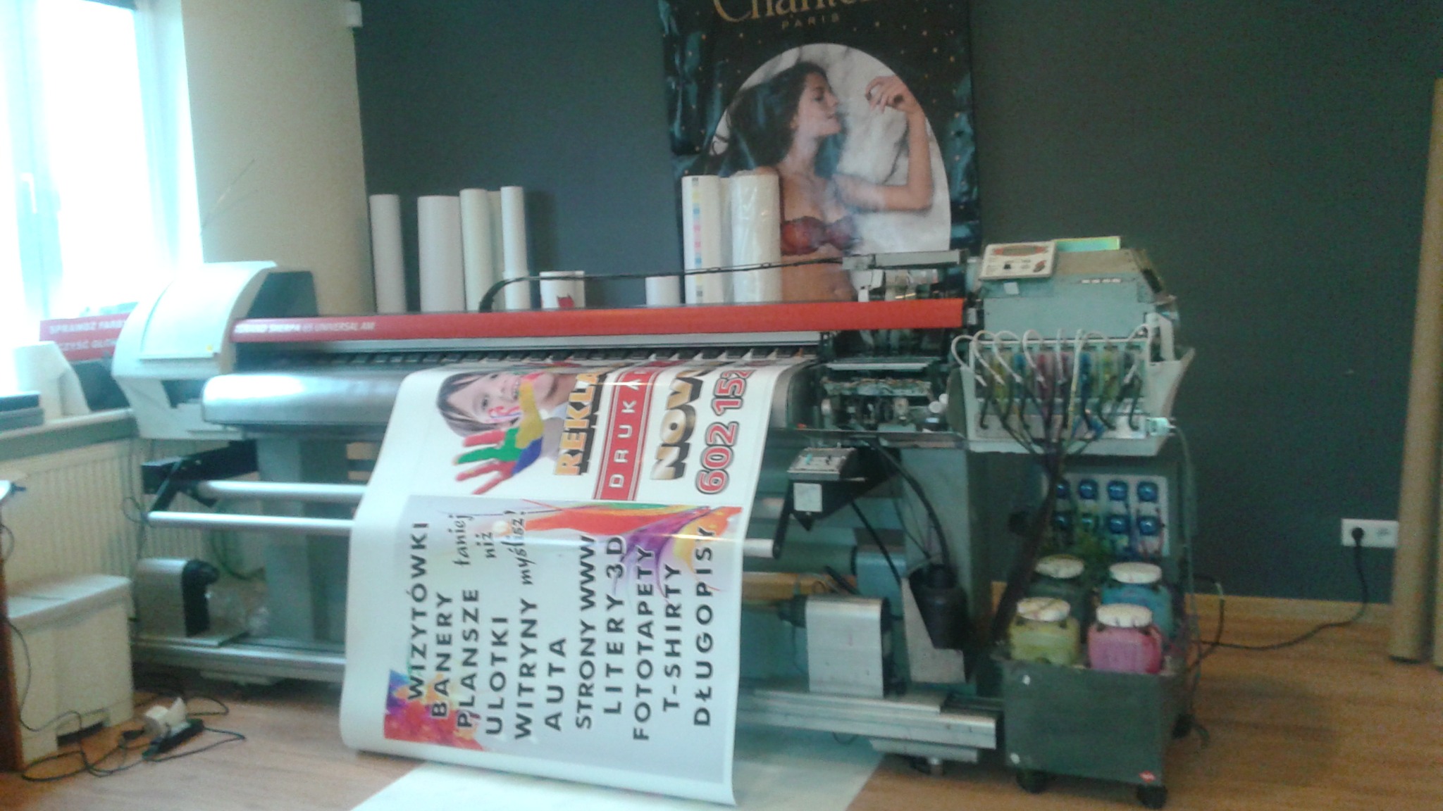 Wielkoformatowa drukarka atramentowa w trakcie drukowania baneru reklamowego z kolorową grafiką, widoczne pojemniki z tuszem i rolki papieru w tle.