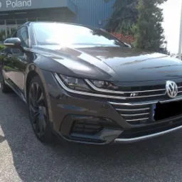 Elegancki, grafitowy Volkswagen Arteon R-Line zaparkowany przed budynkiem z napisem 'O & CO Poland'. Widoczne detale: chromowane listwy, reflektory LED.