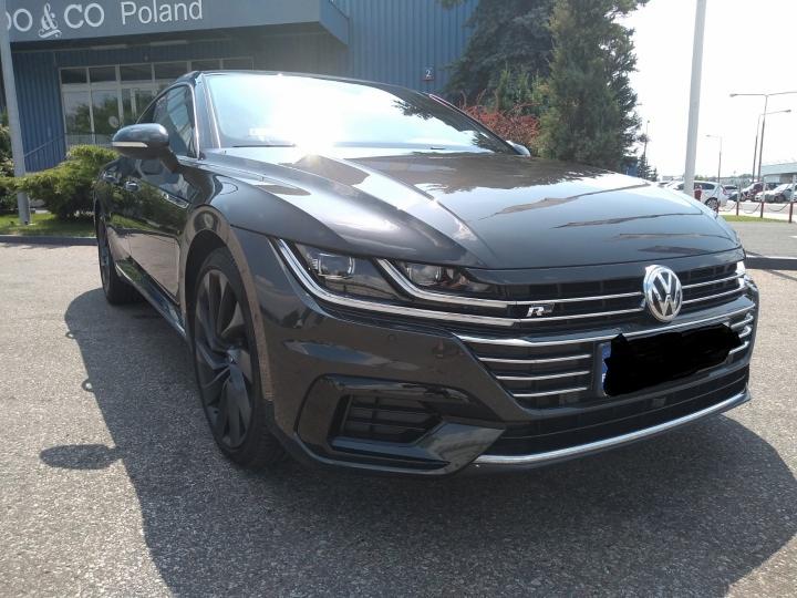 Elegancki, grafitowy Volkswagen Arteon R-Line zaparkowany przed budynkiem z napisem 'O & CO Poland'. Widoczne detale: chromowane listwy, reflektory LED.