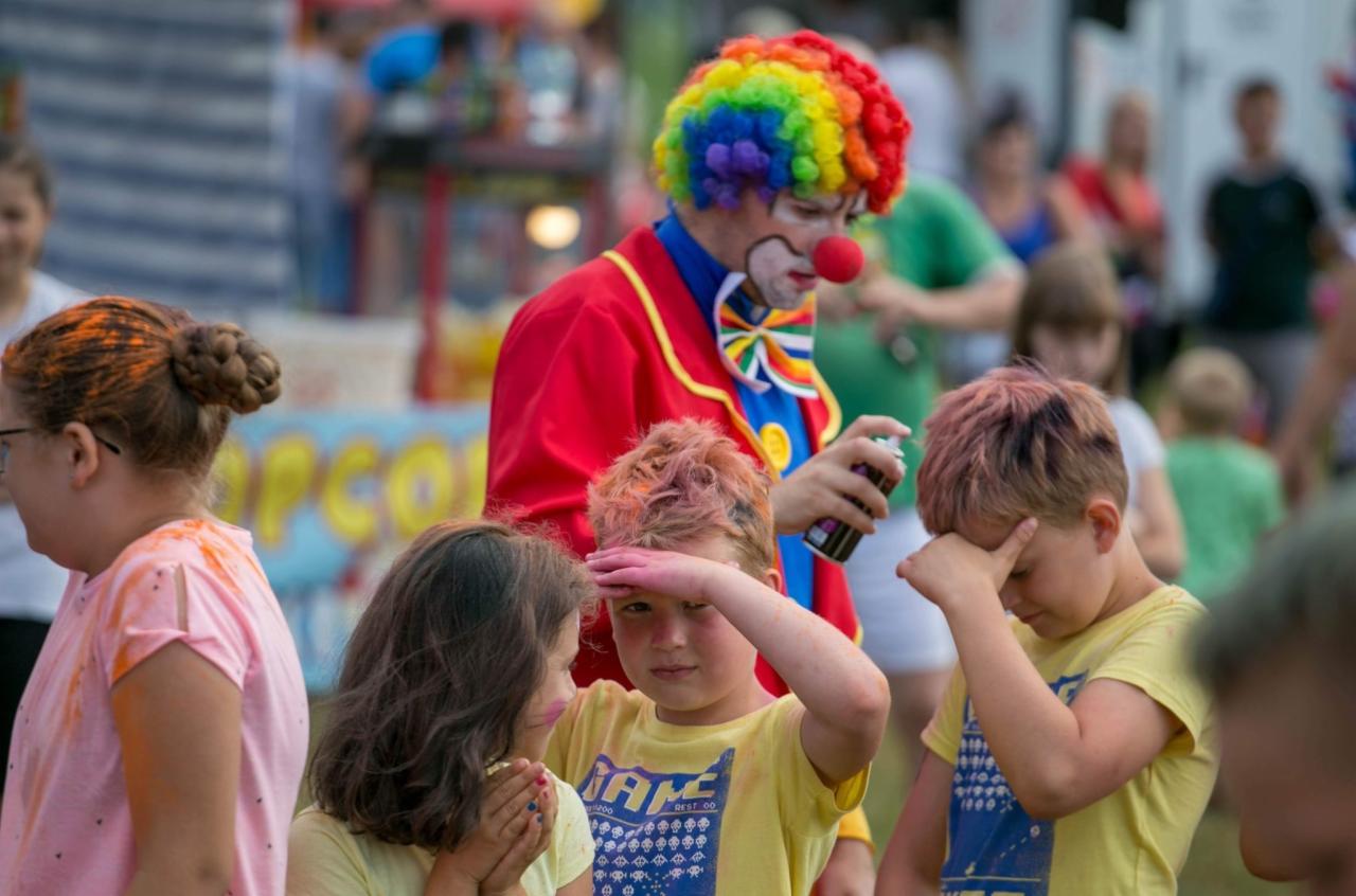 Klaun w czerwonym stroju i tęczowej peruce rozpyla kolorowy spray na włosy dzieciom podczas imprezy plenerowej.
