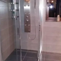 Nowoczesna kabina prysznicowa z panelem hydromasażu, szklane drzwi przesuwne, ciemne płytki na podłodze i ścianach, okno z drewnianą ramą w tle.