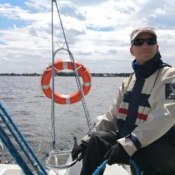 Mężczyzna w kurtce żeglarskiej siedzi na jachcie. W tle woda, niebo z chmurami i brzeg. Na pierwszym planie koło ratunkowe i liny. Słoneczny dzień.