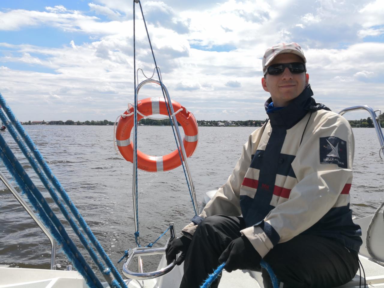 Mężczyzna w kurtce żeglarskiej siedzi na jachcie. W tle woda, niebo z chmurami i brzeg. Na pierwszym planie koło ratunkowe i liny. Słoneczny dzień.