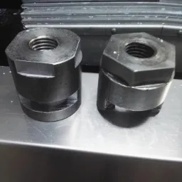 Dwa metalowe elementy z gwintem wewnętrznym i sześciokątną podstawą, leżące na metalowej powierzchni, widoczne ślady obróbki.