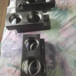 Surowe, metalowe bloki z precyzyjnie wyciętymi okrągłymi otworami, ułożone na wzorzystej tkaninie, widoczne ślady obr&oacute;bki.