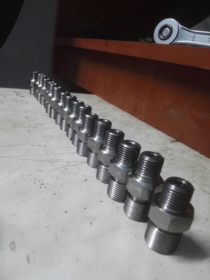 Szereg identycznych, precyzyjnie obrobionych metalowych złączek gwintowanych ułożonych ukośnie na brudnym blacie, klucz oczkowy w tle.