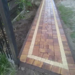 Ułożony chodnik z brązowo-żółtej kostki z obramowaniem z jasnego żwiru, widoczny fragment metalowego ogrodzenia i częściowo wózek budowlany w tle.