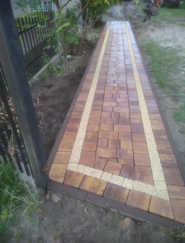 Ułożony chodnik z brązowo-żółtej kostki z obramowaniem z jasnego żwiru, widoczny fragment metalowego ogrodzenia i częściowo wózek budowlany w tle.