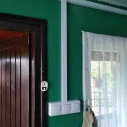 Biała listwa elektroinstalacyjna z zamontowanym potrójnym włącznikiem światła na zielonej ścianie, widoczna drewniana futryna drzwi i okno z firanką.