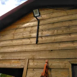 Zewnętrzny reflektor LED zamontowany na drewnianej ścianie budynku z widocznym przewodem w karbowanej osłonie i uchwytem na narzędzia malarskie poniżej.