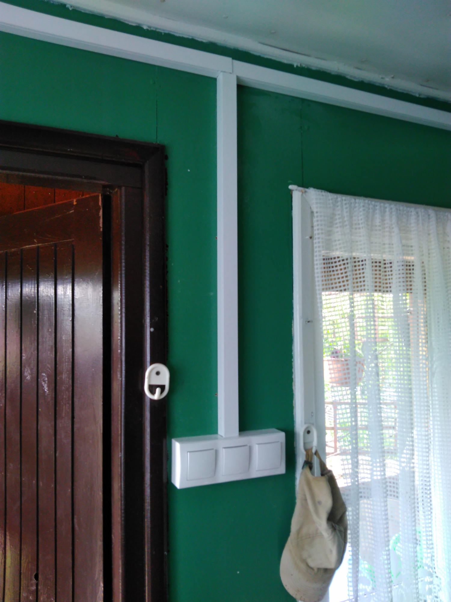 Biała listwa elektroinstalacyjna z zamontowanym potrójnym włącznikiem światła na zielonej ścianie, widoczna drewniana futryna drzwi i okno z firanką.