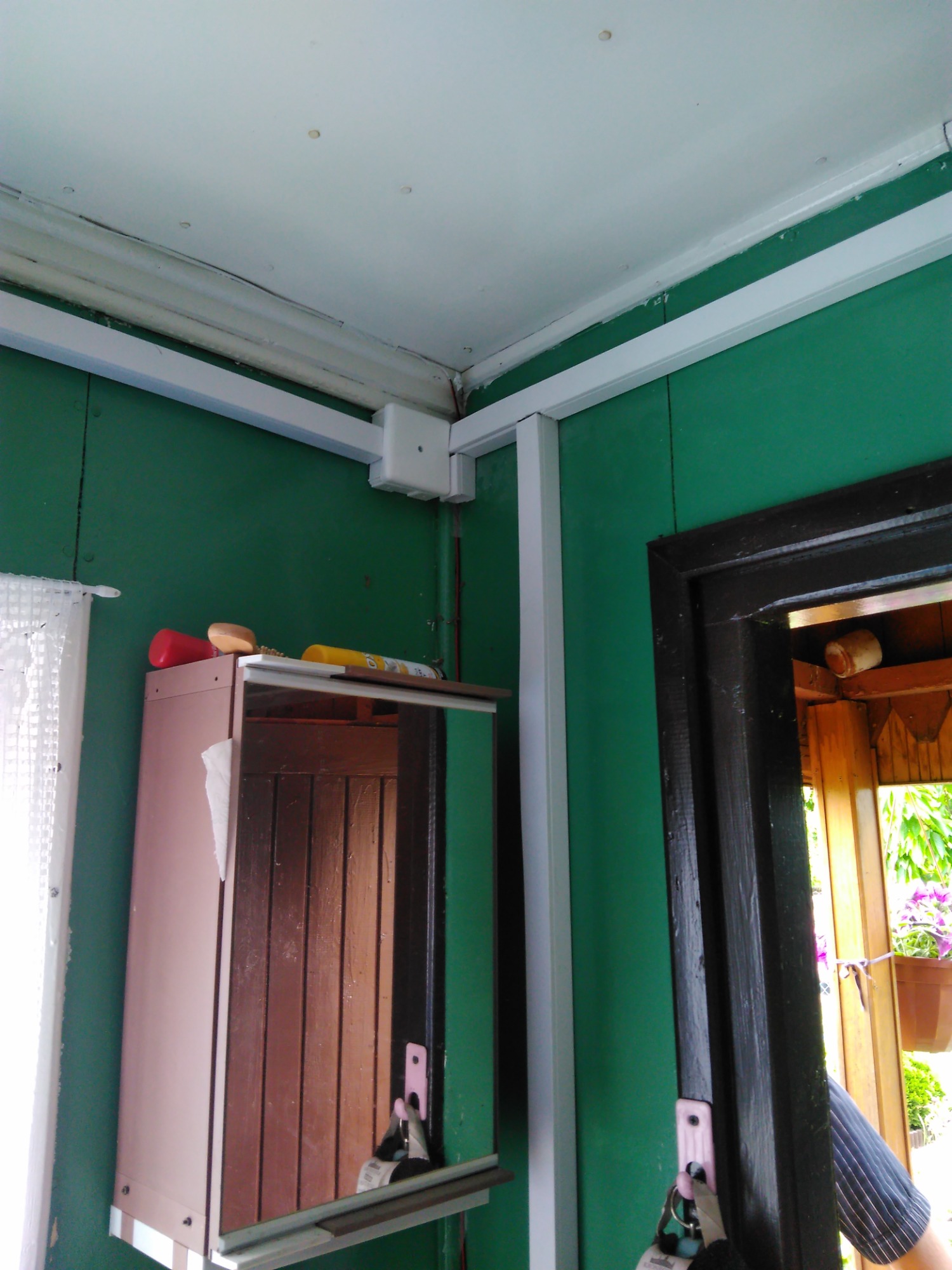 Białe listwy maskujące kable elektryczne biegnące wzdłuż zielonej ściany i sufitu, z puszką łączeniową w rogu, nad szafką z lustrem i wejściem do pomieszczenia.