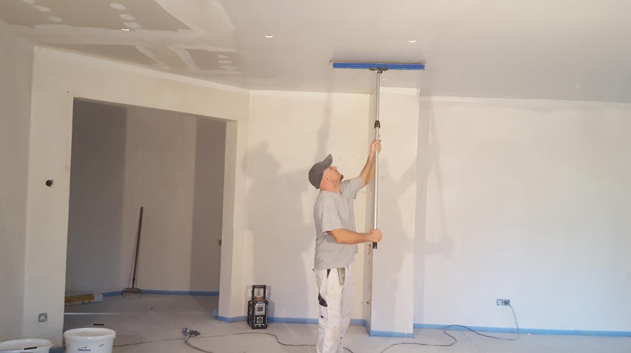 Pracownik w Kluczborku wygładza sufit długą szpachlą na teleskopowym kiju, widoczne ślady po łączeniach płyt gipsowych.