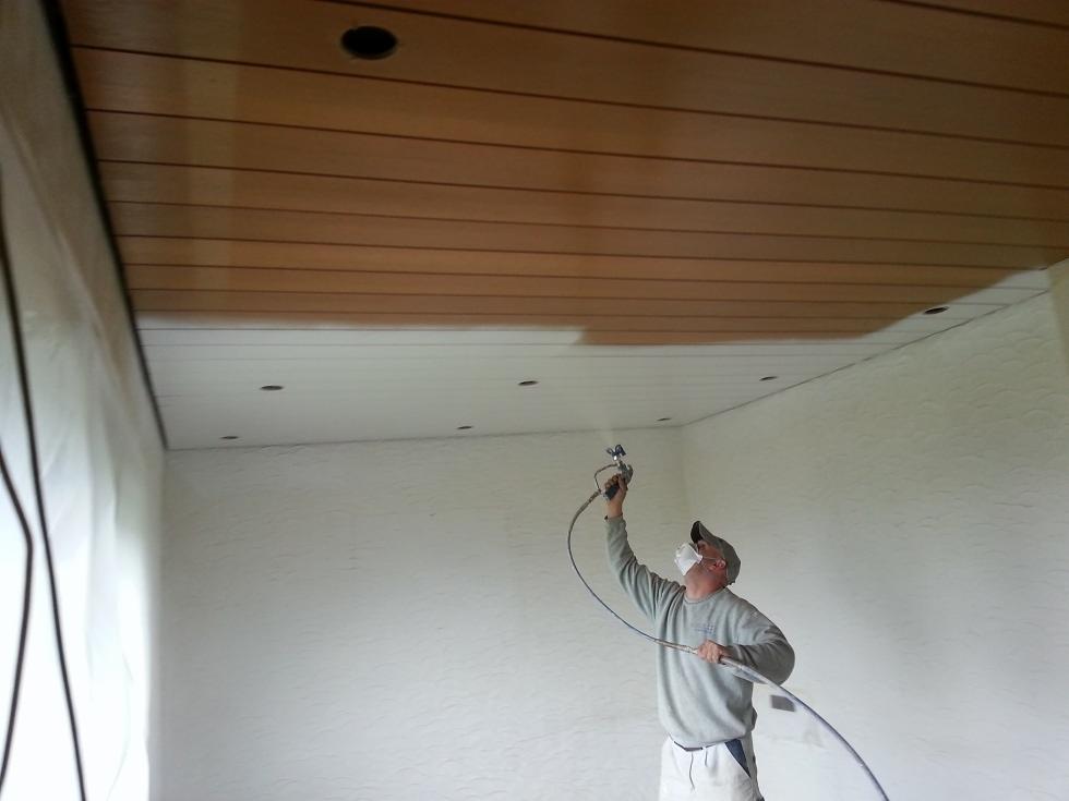 Pracownik w masce maluje natryskowo sufit z drewnianą boazerią, widoczna granica pomalowanej i niepomalowanej powierzchni.