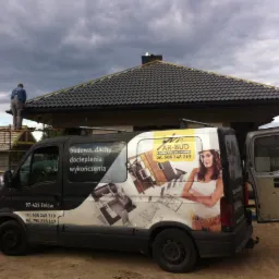 Nowy dom w budowie z ciemną dachówką, widoczny zza firmowego busa z reklamą usług budowlanych i numerem telefonu, na tle pochmurnego nieba. Jeden z pracowników na dachu.