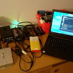 Diagnostyka ECU samochodu BMW za pomocą laptopa Lenovo i interfejsu KESS Alientech, widoczne podłączenie do sterownika silnika, zasilacz i dodatkowe kable na drewnianym biurku.