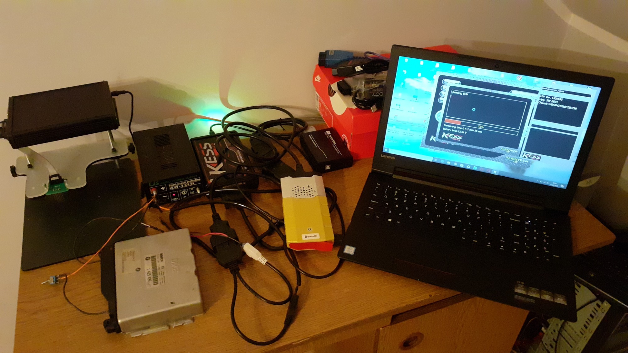 Diagnostyka ECU samochodu BMW za pomocą laptopa Lenovo i interfejsu KESS Alientech, widoczne podłączenie do sterownika silnika, zasilacz i dodatkowe kable na drewnianym biurku.