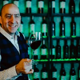 Uśmiechnięty mężczyzna w kamizelce w kratę trzyma kieliszek czerwonego wina na tle regału z podświetlonymi butelkami.