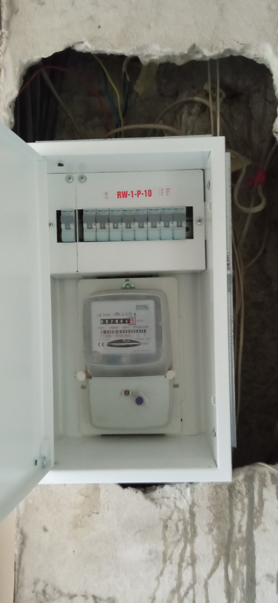 Otwarta, biała skrzynka elektryczna z zamontowanym licznikiem zużycia energii i bezpiecznikami, widoczna na tle surowej ściany z wystającymi przewodami.