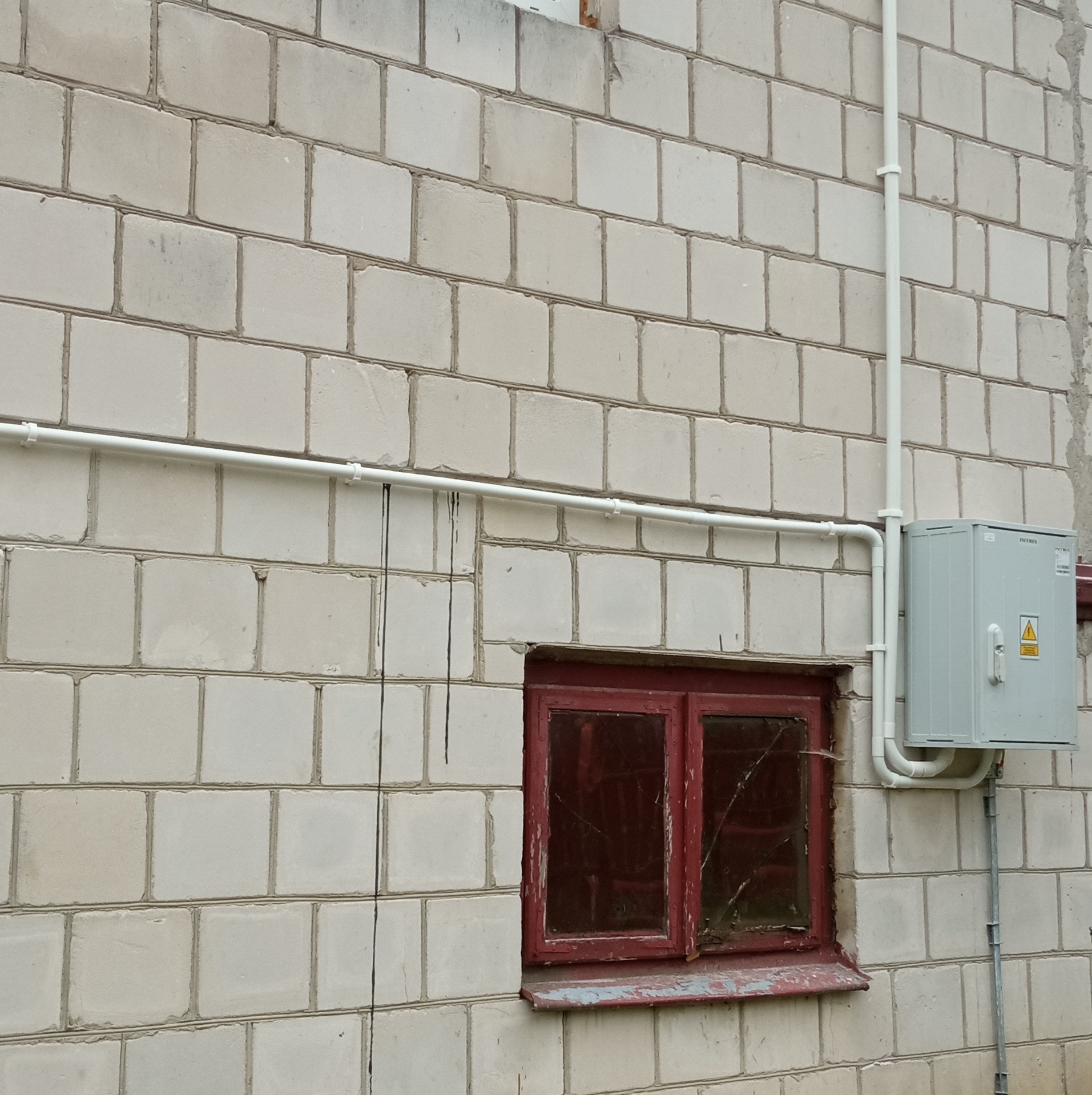 Szara skrzynka elektryczna zamontowana na ścianie z białych bloczków, z widocznymi białymi rurami instalacyjnymi i fragmentem starego, czerwonego okna.