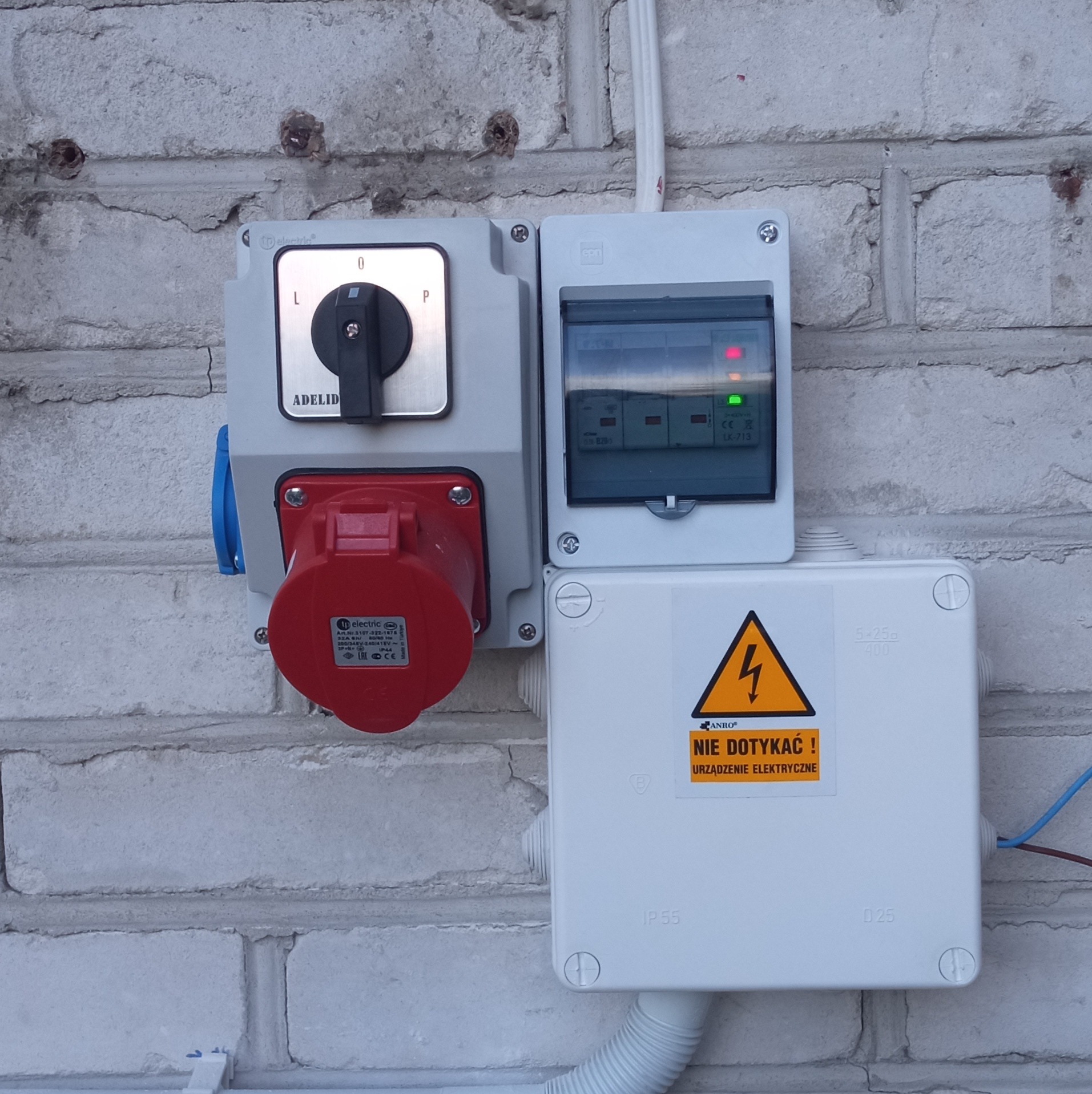 Zewnętrzna skrzynka elektryczna z wyłącznikiem, gniazdem przemysłowym i zabezpieczeniami, zamontowana na ceglanej ścianie. Widoczna tabliczka ostrzegawcza 'Nie dotykać! Urządzenie elektryczne'.