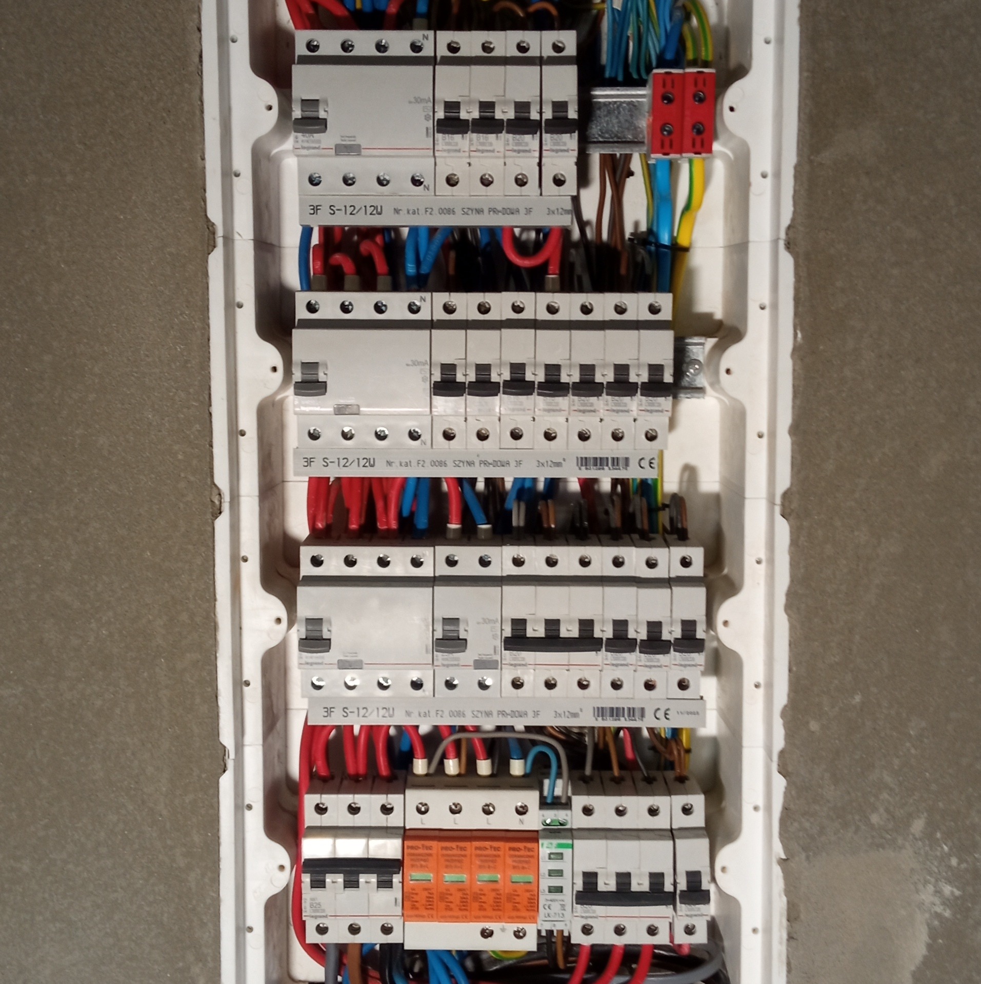 Rozdzielnica elektryczna z zainstalowanymi bezpiecznikami i przewodami w kolorach niebieskim, czerwonym i żółto-zielonym, zamontowana w ścianie z surowego betonu.