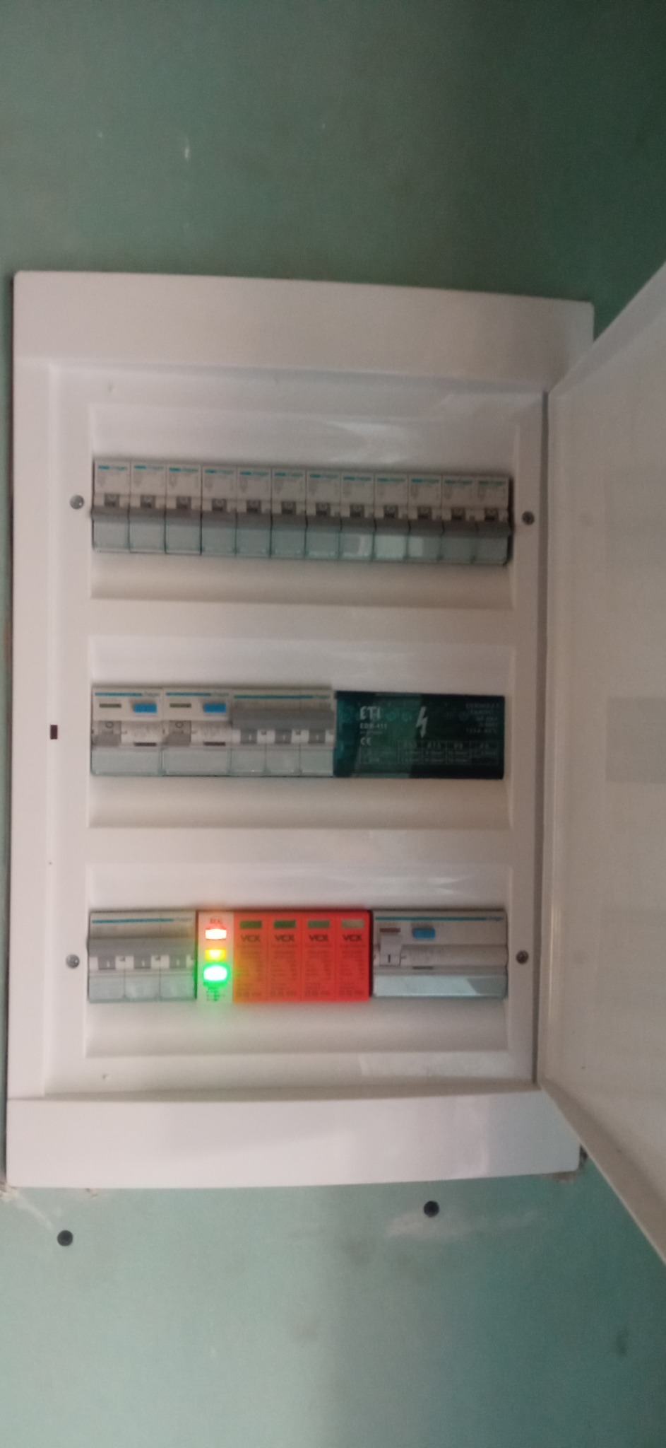 Otwarta skrzynka elektryczna z wyłącznikami, w dolnym rzędzie tester napięcia VOX z zapalonymi diodami, zamontowana na zielonej ścianie.